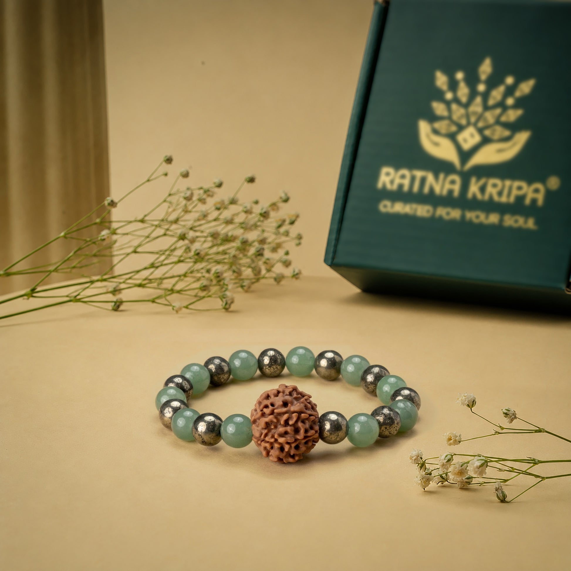Bracelet with Pyrite and Green Aventurine beads band a 7 Mukhi Rudraksha bead on a beige surface with a 'Ratna Kripa' box in the background.
