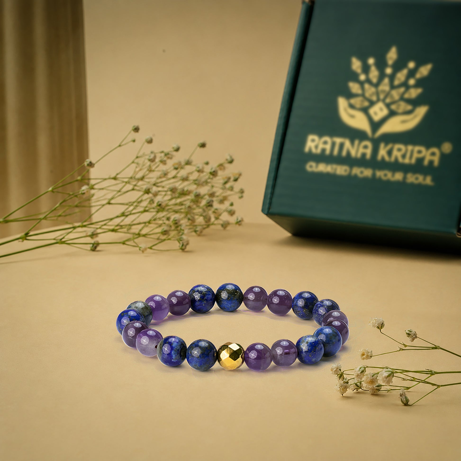 Blue beaded bracelet with a gold bead on a beige surface next to a green box labeled 'Ratna Kripa'.