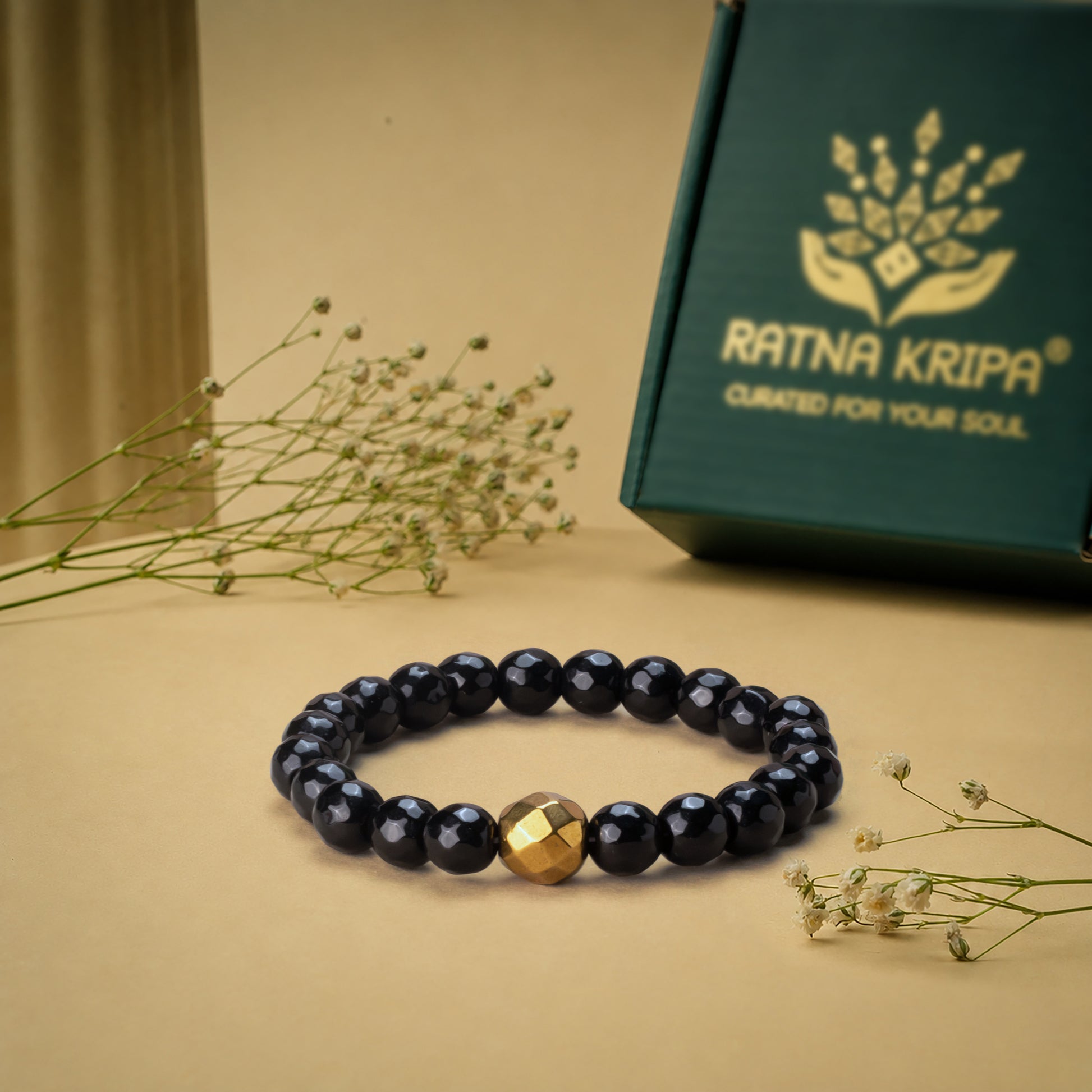 Black Tourmaline Diamond Cut Bracelet with a golden hematite bead on a beige surface, next to a green box with 'Ratna Kripa' branding.