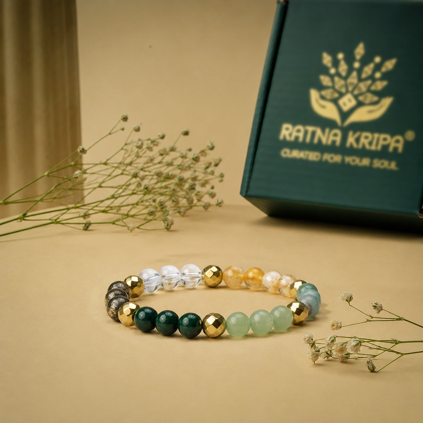 Beaded bracelet with green, gold, and clear beads on a beige surface with a 'Ratna Kripa' box in the background.