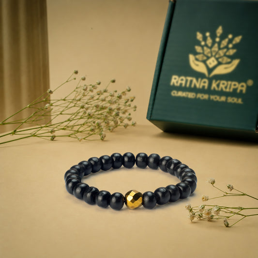 Black Karungali beaded bracelet with a golden hematite accent on a beige surface, next to a green box with 'Ratna Kripa' branding.