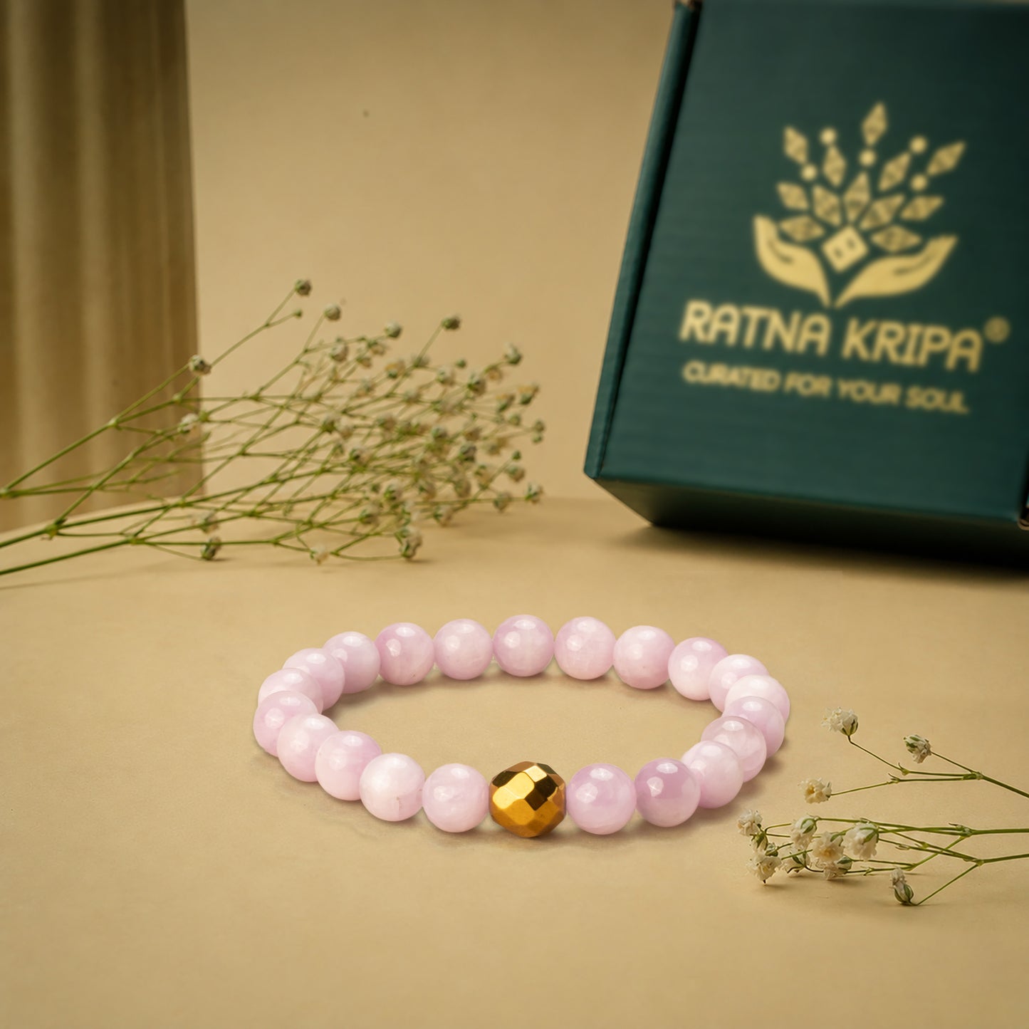 Pink-coloured kunzite bead bracelet with a golden hematite on a beige surface, next to a green box with 'Ratna Kripa' branding.