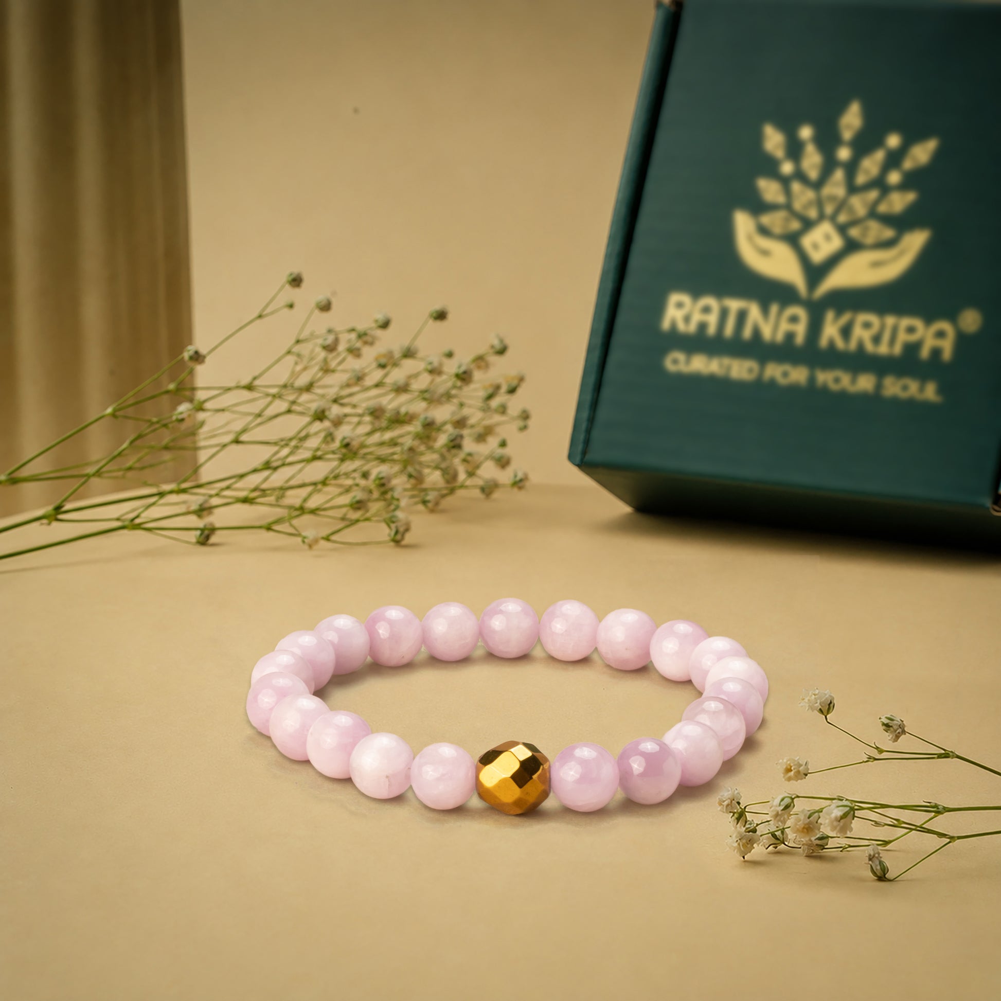 Pink-coloured kunzite bead bracelet with a golden hematite on a beige surface, next to a green box with 'Ratna Kripa' branding.