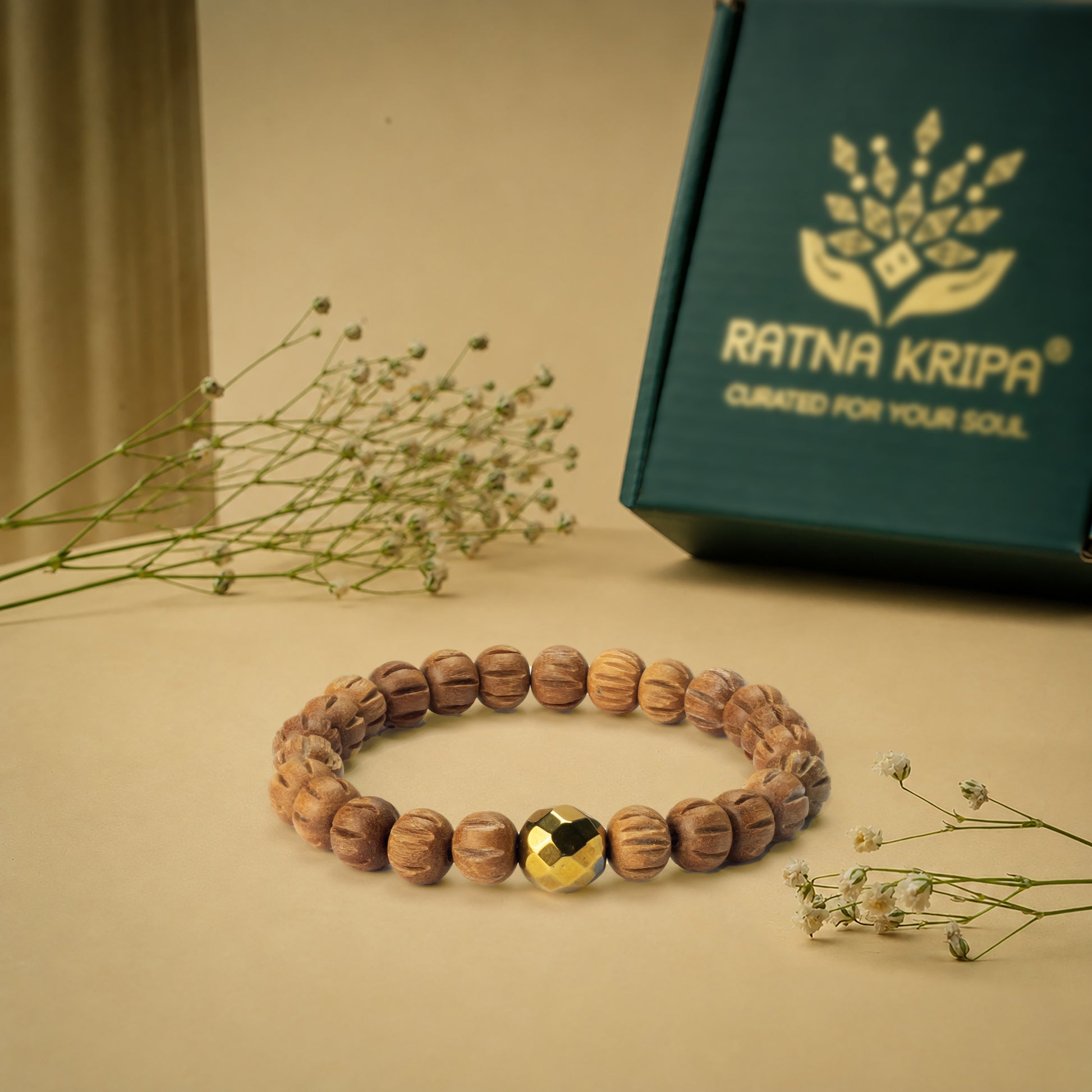 Wooden bracelet with a golden hematite bead on a beige surface next to a green box with 'Ratna Kripa' branding.