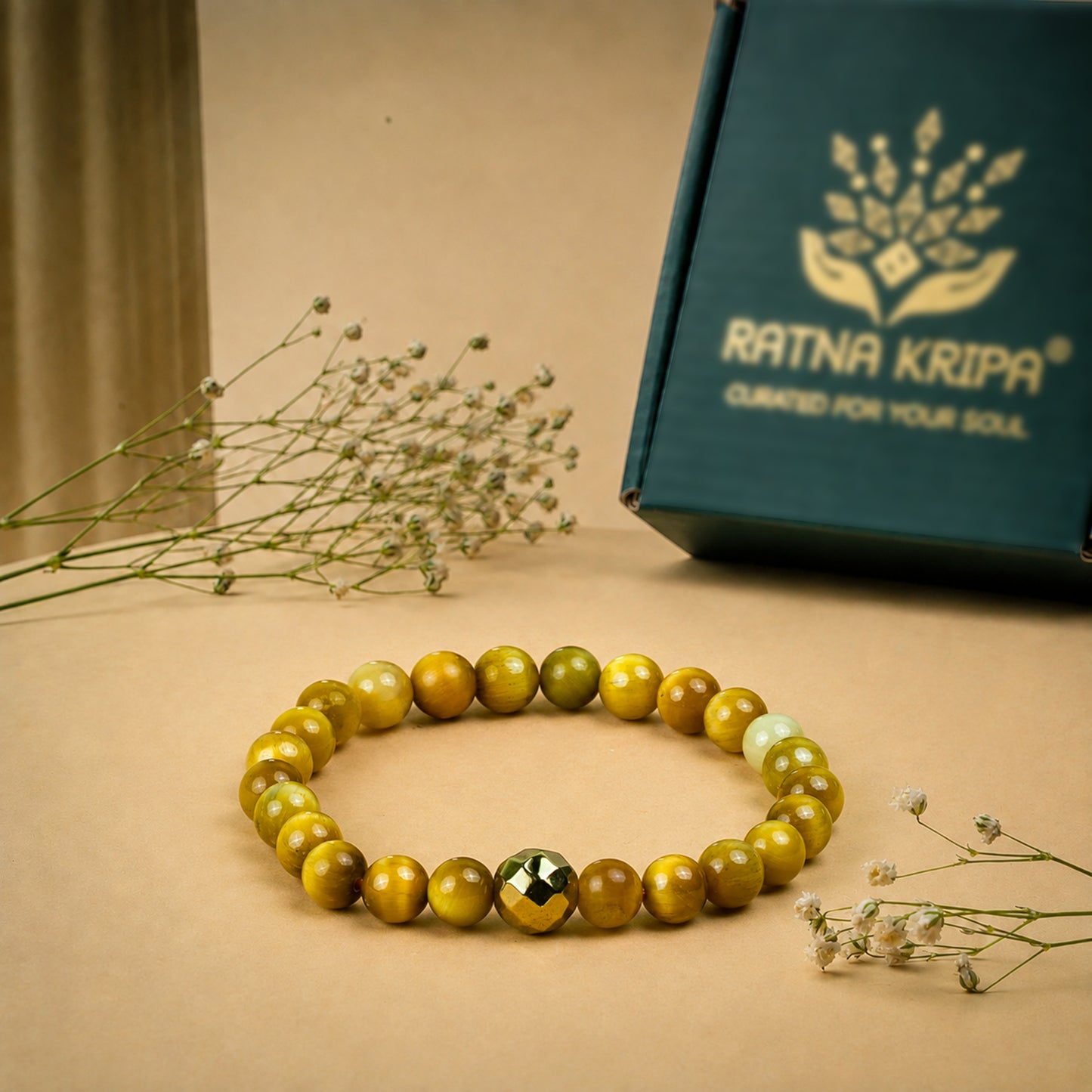 A beaded stretch bracelet made of polished golden-yellow and olive-green round beads with a faceted metallic gold accent bead, displayed on a beige surface with dried baby's breath flowers and a dark teal gift box branded RATNA KRIPA in the background.